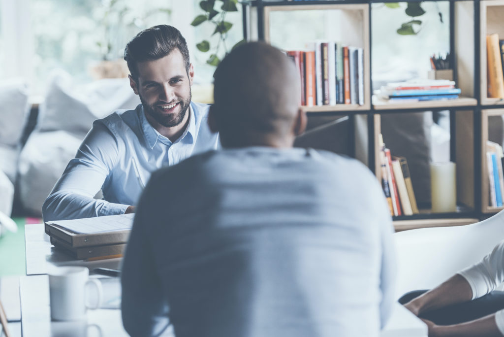 Two young men in smart casual wear sitting at the office desk together while one of them smiling